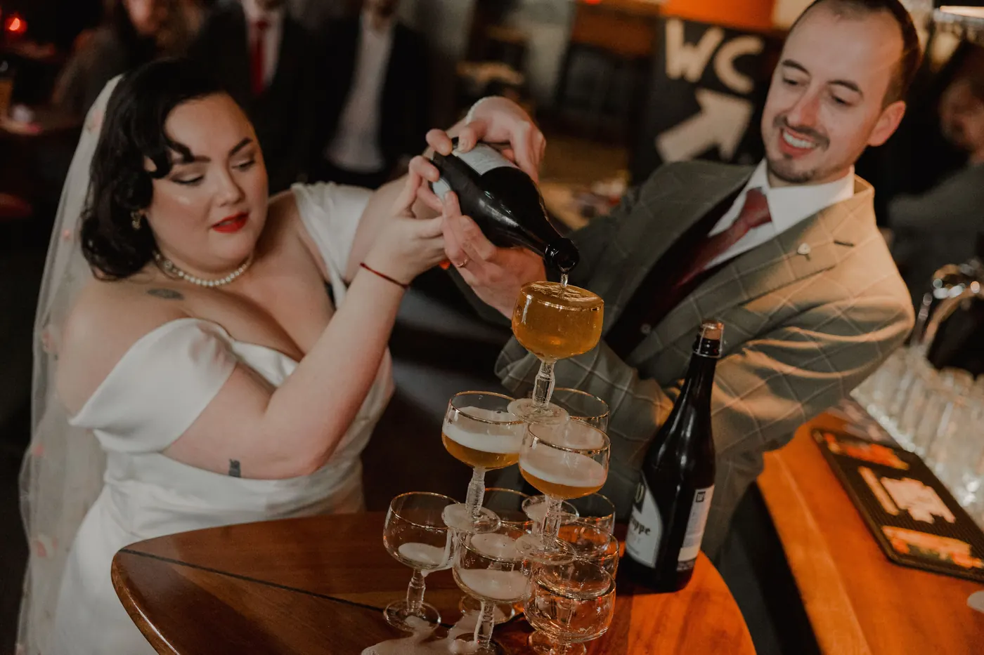 Bruid en bruidegom schenken samen champagne in een toren van coupes op een houten bar